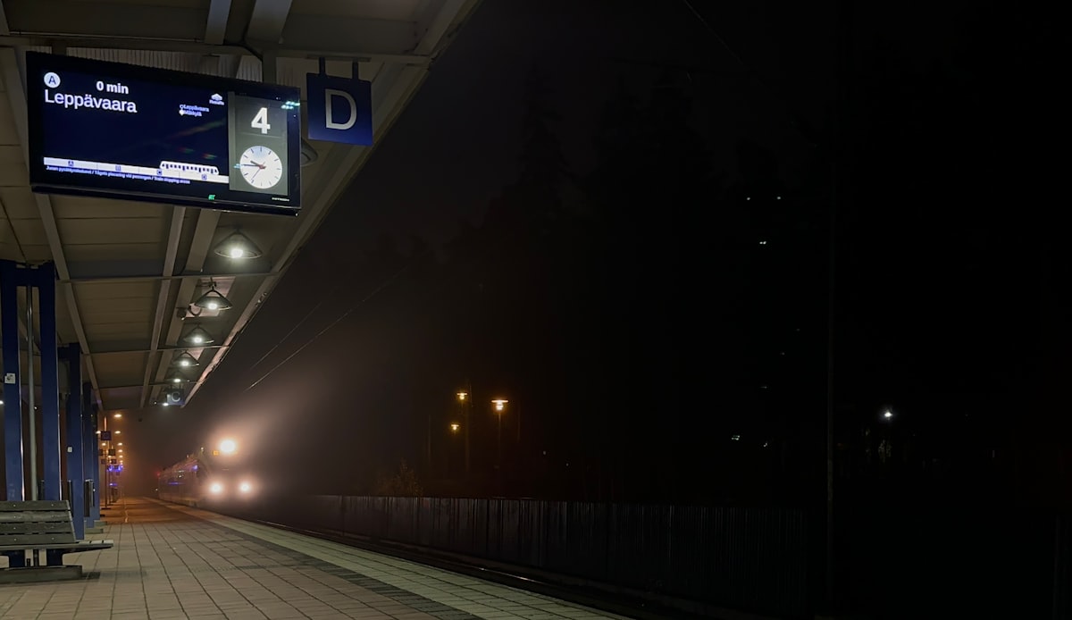 A train approaching an empty railway platform at night in fog, its headlights lit beneath a departure board, no identifiable people and no readable commercial brands or logos, for the piece on Europe's 2026 night-train map