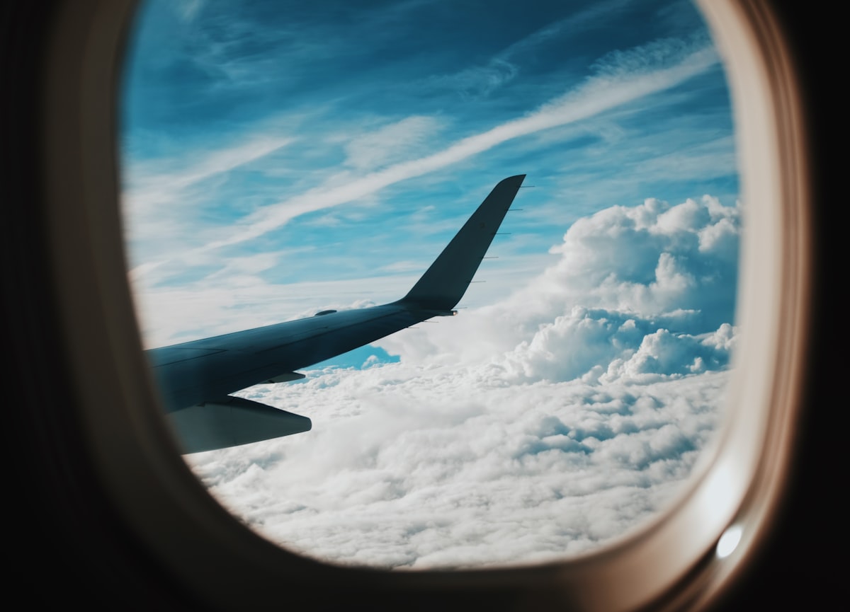 The wing of an airliner seen from a passenger window above the clouds, no identifiable people and no readable airline branding, for the piece on how to actually find a cheap flight now
