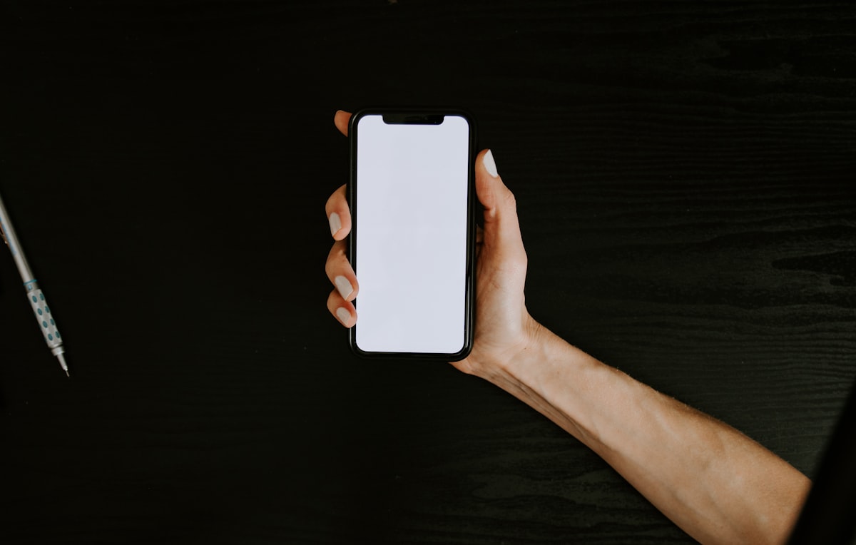A hand holding a smartphone with a blank white screen on a dark desk, no identifiable face and no readable brand or logo, echoing the setup-wizard skip button in the piece on a third of consumers not wanting AI on their devices