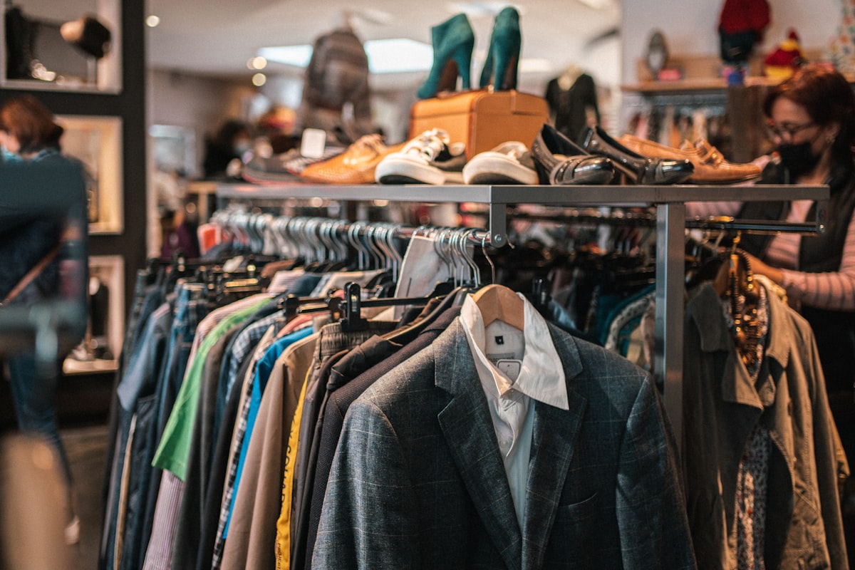 A rack of clothes with shoes displayed above in a store, incidental shoppers out of focus and no readable brand or logo, for the piece on the EU ban on destroying unsold clothes and shoes
