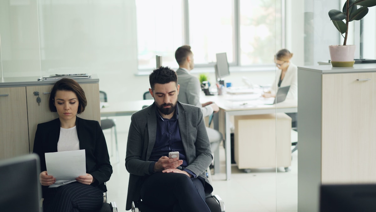 Two job candidates waiting in an office lobby with resumes in hand, for the piece on recent graduates competing for a shrinking number of entry-level openings