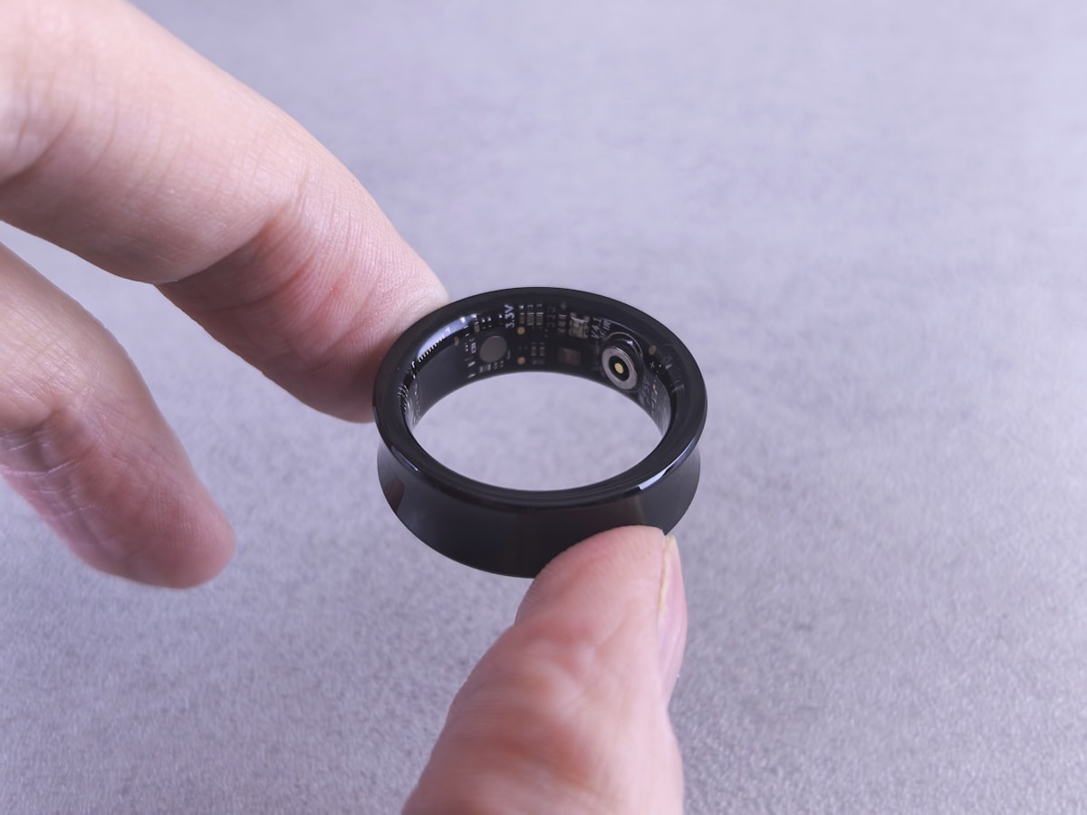 Fingers holding a black smart ring with its internal circuitry visible, no face shown, for the piece on the quiet trade-offs of ring wearables