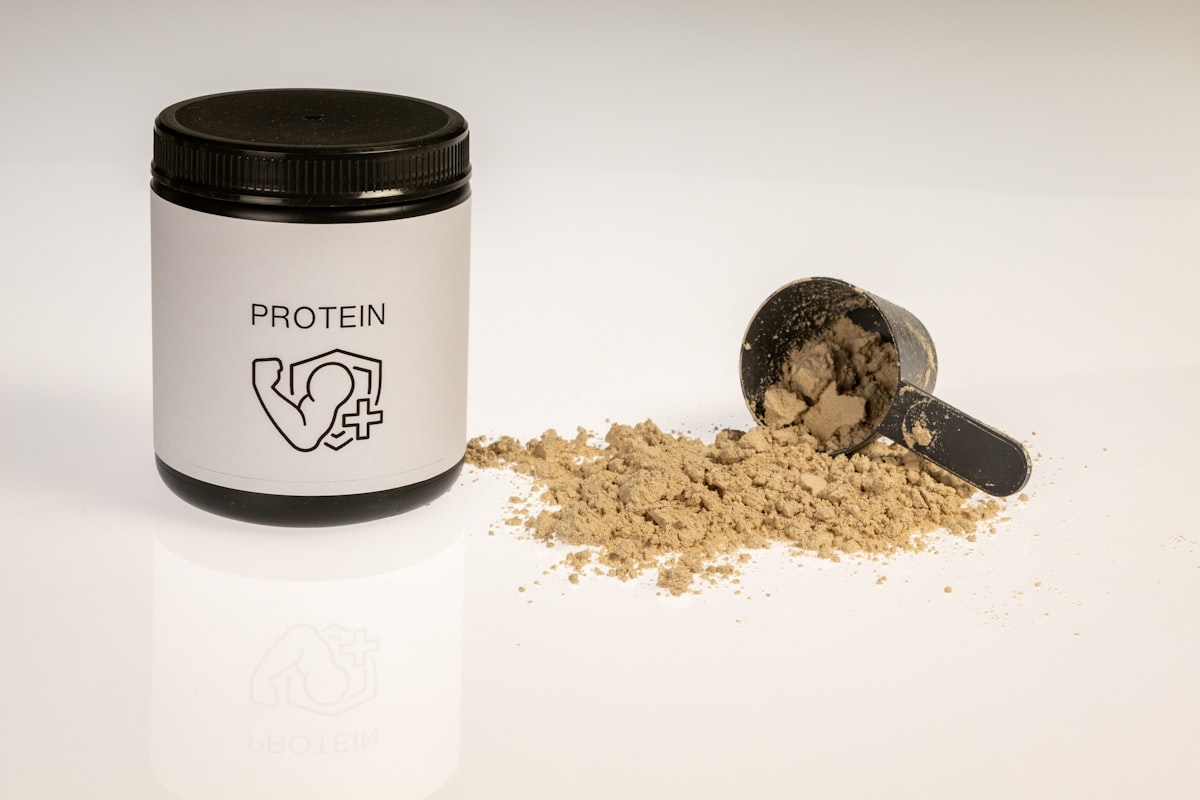 A generic supplement tub labeled only with the word PROTEIN beside a spilled scoop of powder on a white background, no identifiable people and no readable real-brand name or logo, for the piece on protein-timing marketing