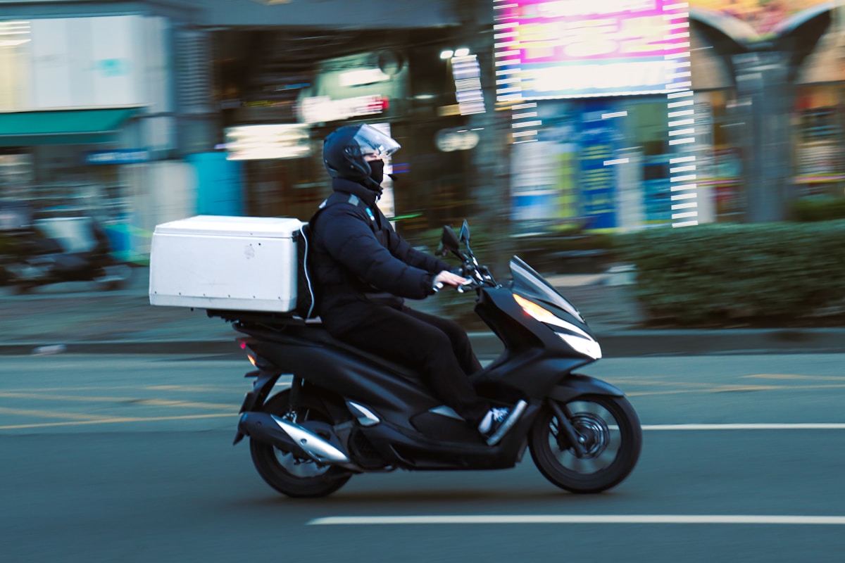 A helmeted delivery courier on a scooter with a plain unbranded white top-box, in motion on a city street, no identifiable face and no readable brand or logo, for the piece on the EU Platform Work Directive and gig-platform couriers