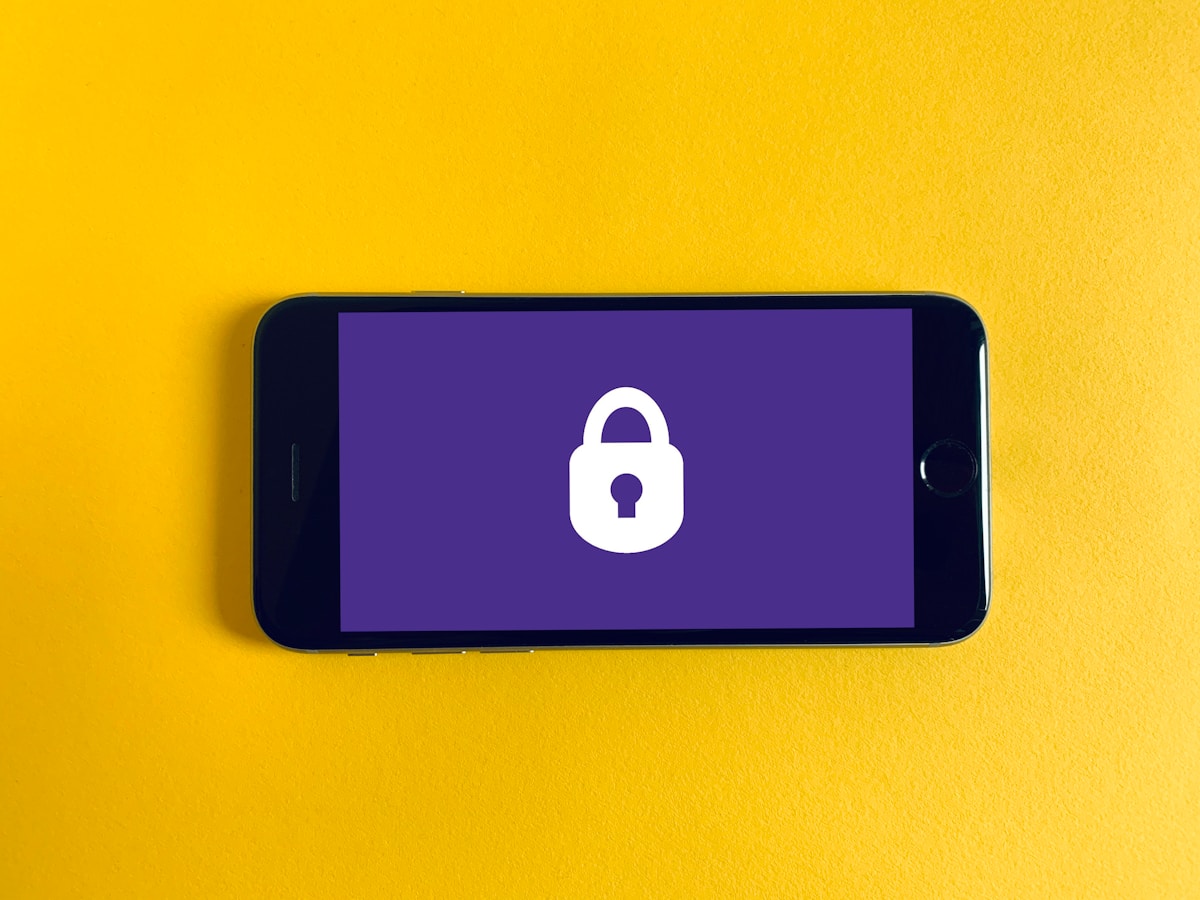 A smartphone lying face-up on a plain background, its screen showing a generic white padlock icon on a purple field, no face, no readable brand or logo, and no real ID document, standing in for the phone-based identity checkpoint at the center of the piece on age-verification laws pulling ID checks into phones and VPNs