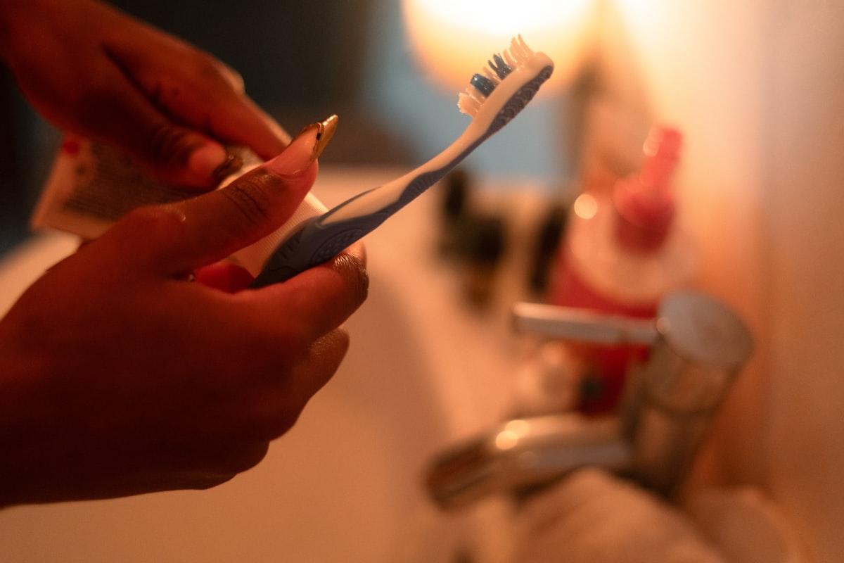 Generic hands squeezing unbranded toothpaste onto a toothbrush at a bathroom sink, no face, no readable brand or logo, for the piece on titanium dioxide's unresolved status in oral cosmetics