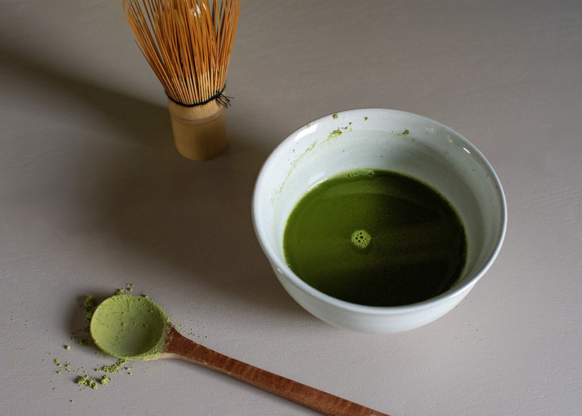 A white bowl of whisked matcha beside a bamboo whisk and a wooden scoop of matcha powder on a neutral surface, no identifiable people and no readable brands or logos, for the piece on the 2025 matcha price spike
