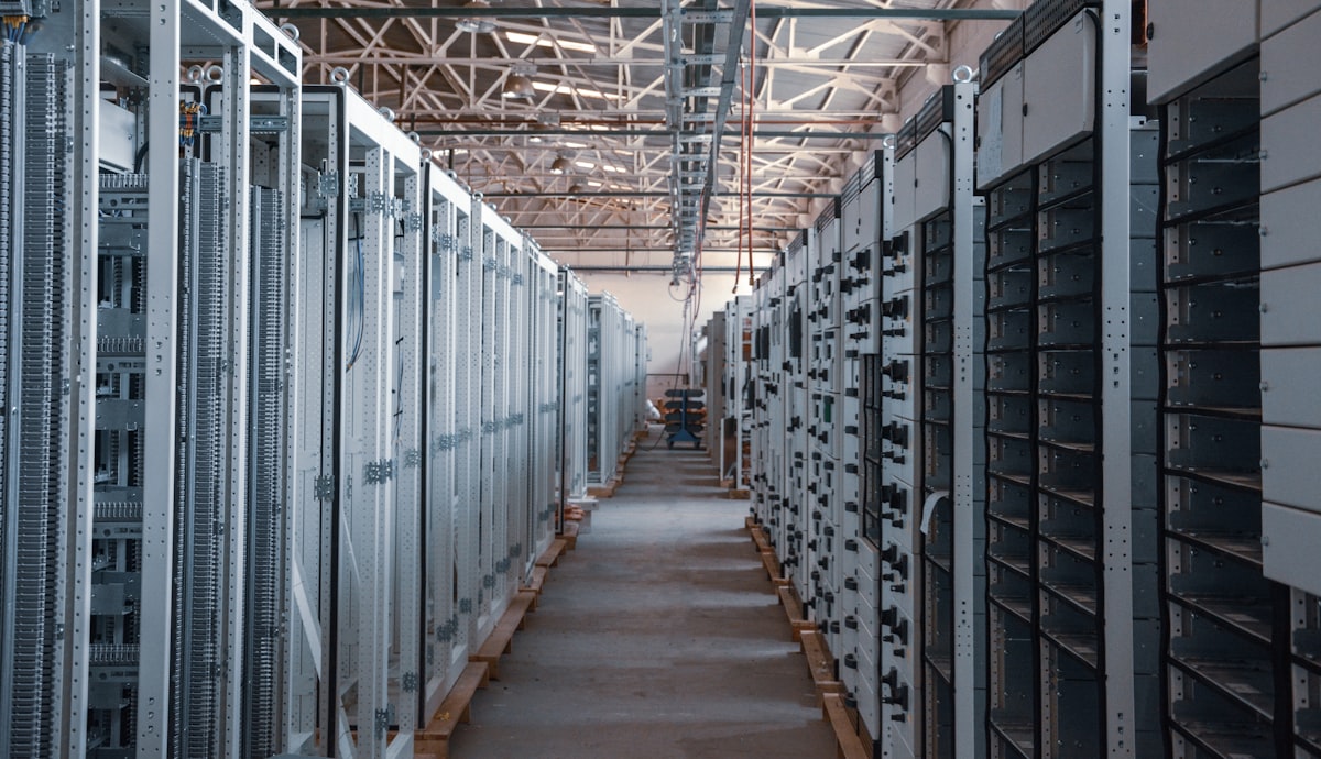 A long aisle between rows of metal server and equipment racks in a data center, no identifiable people and no readable brands or logos, for the piece on whether AI's data-center buildout is a bubble