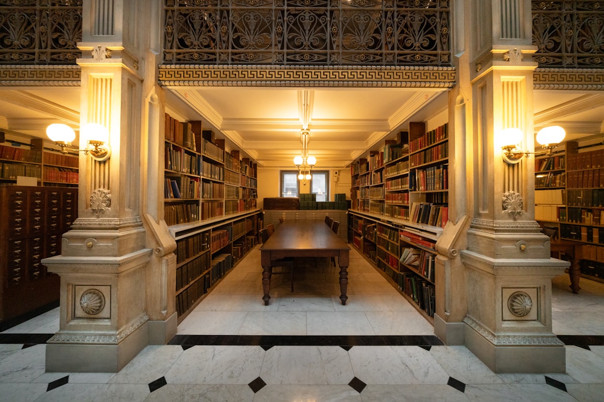 A public library reading room with long tables between tall book stacks and pillars, no identifiable person and no readable brand, illustrating the public library as the third place that asks nothing of you and cannot be priced out