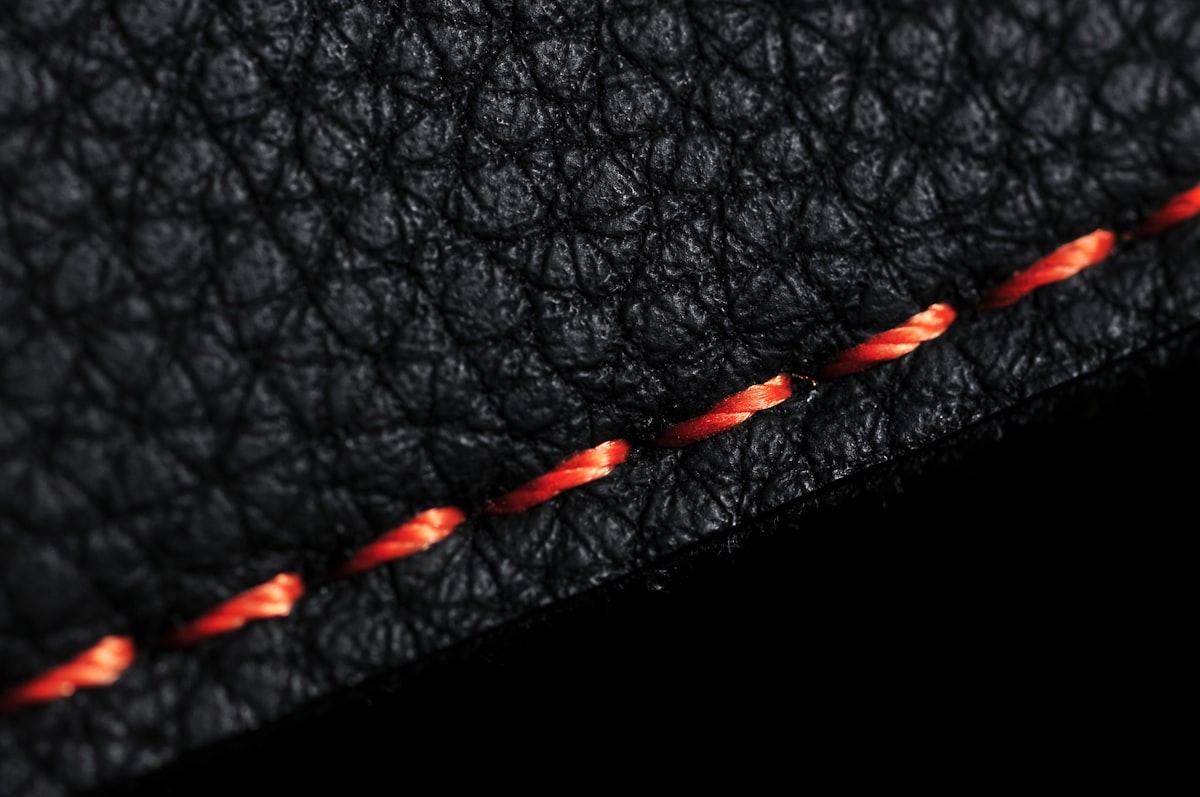 A macro close-up of a plain black pebbled-leather seam with a single line of orange contrast stitching, no hardware, no identifiable person, and no readable brand, logo, or monogram, for the paragraph on Entrupy reading fabric, stitching, and metal under a microscope attachment
