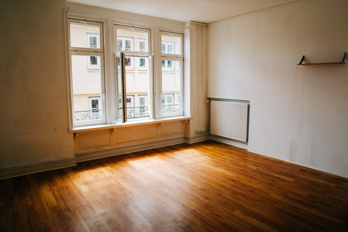 An empty apartment room with a wooden floor, bare walls, and daylight through a tall window, no identifiable people and no readable brands or logos, for the piece on stretched emerging adulthood and the cost of housing