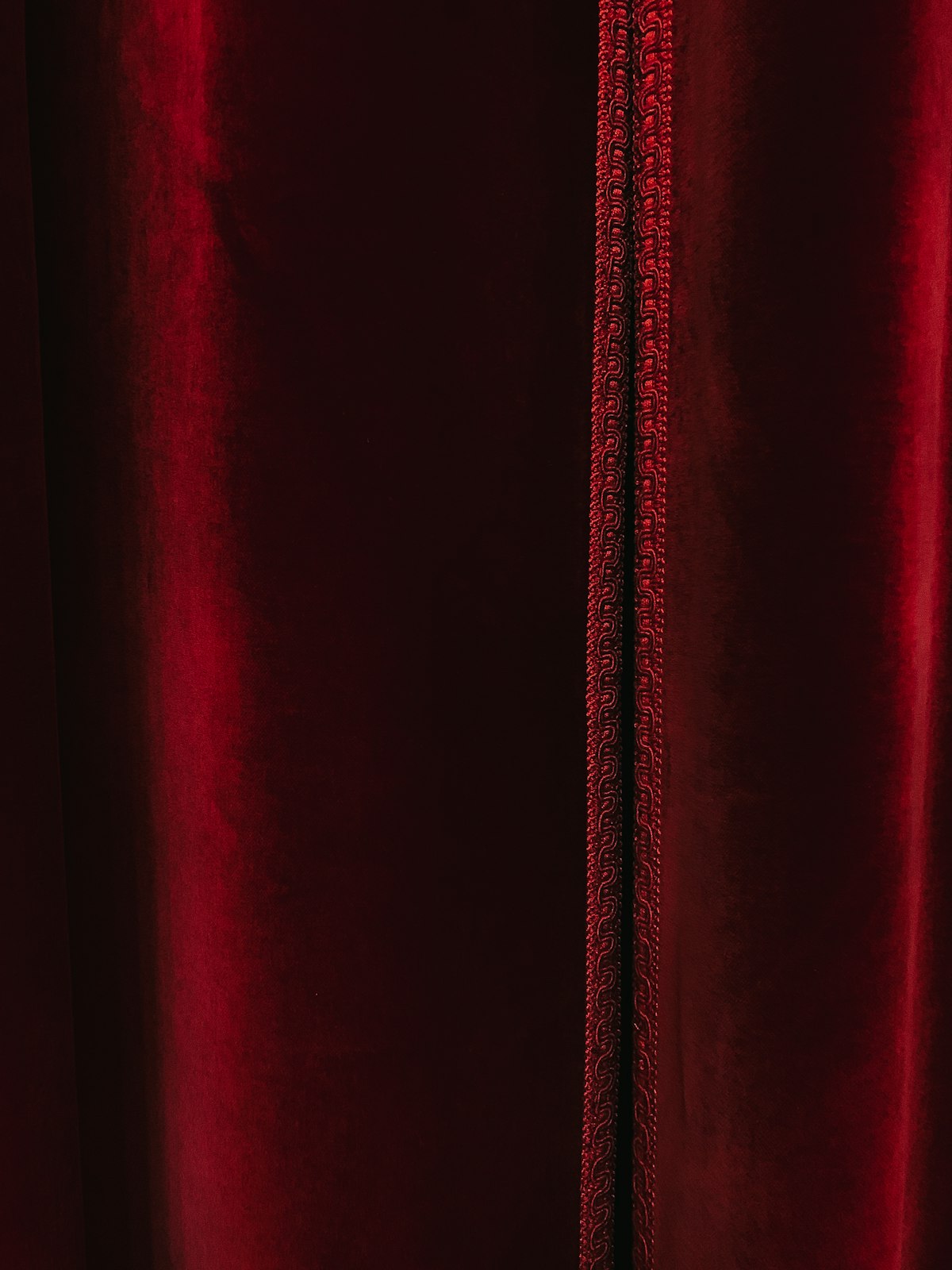 A close detail of deep red velvet with a raised braided cord trim running vertically down a seam, no identifiable people and no readable brand or logo, illustrating the braided frogging and seams to inspect on a secondhand velvet jacket