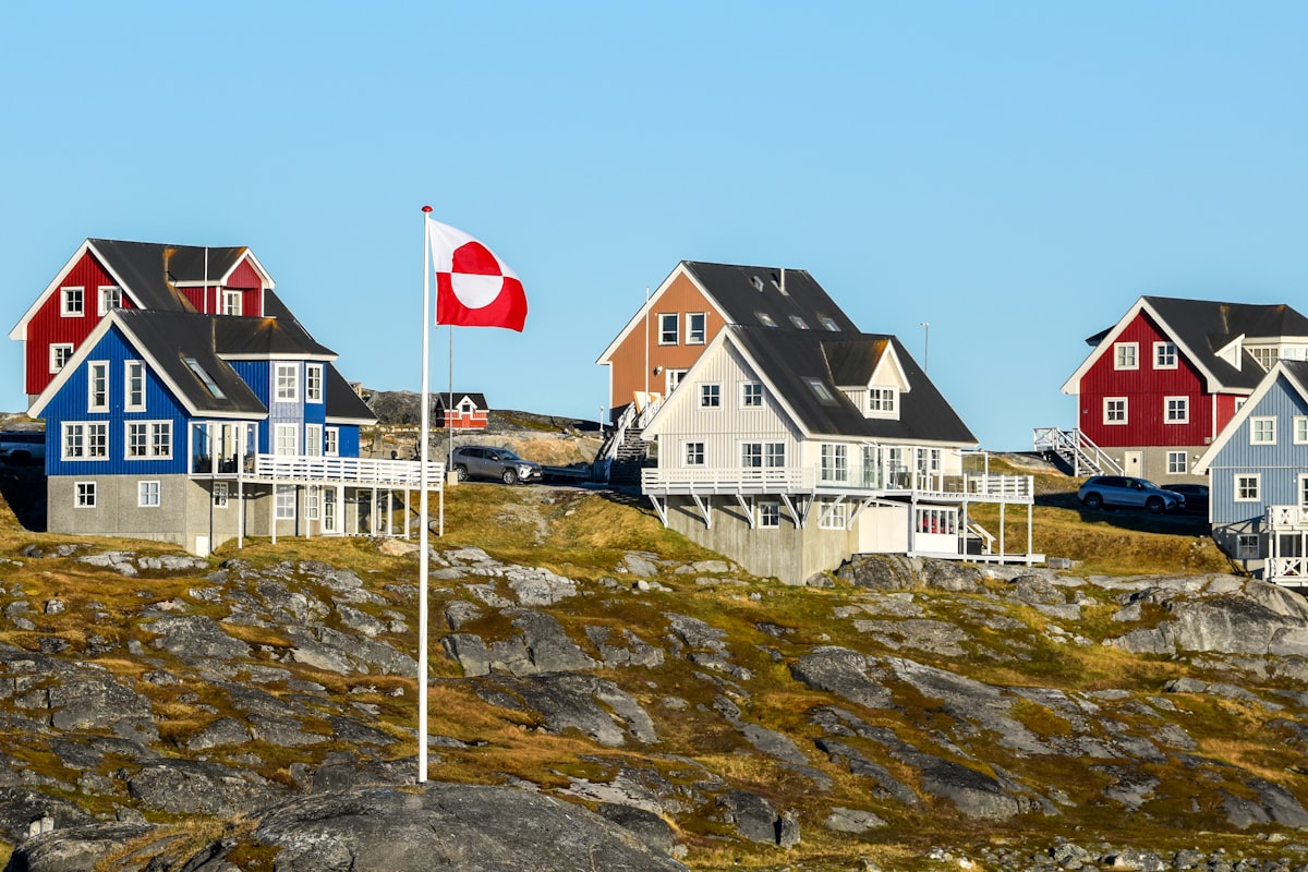 Brightly painted wooden houses on bare rock beneath a clear sky with a Greenlandic flag flying in front, no identifiable people and no readable commercial brands or logos, for the piece on Greenland's tourism law